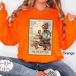 May include: Orange sweatshirt featuring a graphic of a skeleton holding a coffee cup and hockey stick. The graphic is in a vintage style and says "The Hockey Mom".