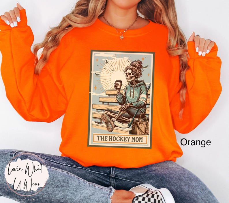May include: Orange sweatshirt featuring a graphic of a skeleton holding a coffee cup and hockey stick. The graphic is in a vintage style and says "The Hockey Mom".