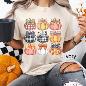 Peut inclure: T-shirt ivoire avec une grille de neuf citrouilles aux motifs et n&oelig;uds vari&eacute;s. Les citrouilles sont orange, rose, jaune et blanches, avec des n&oelig;uds &agrave; carreaux, &agrave; pois et unis. Le mot "Ivory" est visible.