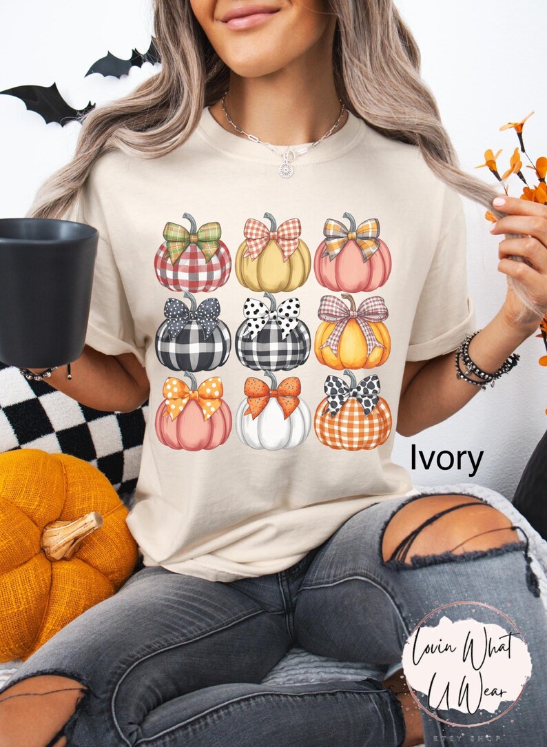 Peut inclure: T-shirt ivoire avec une grille de neuf citrouilles aux motifs et n&oelig;uds vari&eacute;s. Les citrouilles sont orange, rose, jaune et blanches, avec des n&oelig;uds &agrave; carreaux, &agrave; pois et unis. Le mot "Ivory" est visible.