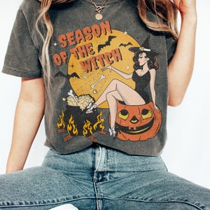 Puede incluir: Una camiseta gris oscuro con una ilustraci&oacute;n vintage de una bruja sentada en una calabaza de Halloween. El texto "Season of the Witch" est&aacute; impreso en la camiseta.