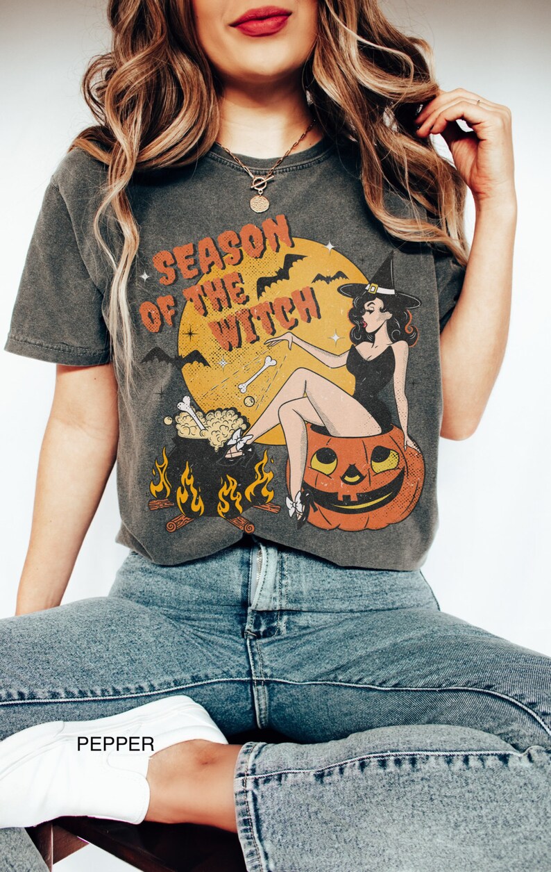 Puede incluir: Una camiseta gris oscuro con una ilustraci&oacute;n vintage de una bruja sentada en una calabaza de Halloween. El texto "Season of the Witch" est&aacute; impreso en la camiseta.
