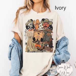 May include: Ivory-colored t-shirt with a Halloween-themed graphic. The design includes a skull, cactus, pumpkin, and other spooky elements. Text on the shirt reads "October 31" and "Halloween Never Die."