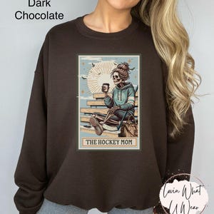 May include: A dark chocolate brown sweatshirt featuring a graphic of a skeleton sitting on a bench with a coffee cup and hockey stick. The graphic is in a vintage style with a light blue background and the text "The Hockey Mom".