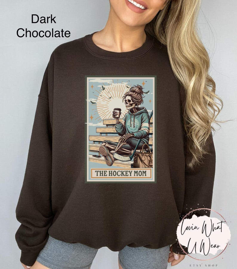 May include: A dark chocolate brown sweatshirt featuring a graphic of a skeleton sitting on a bench with a coffee cup and hockey stick. The graphic is in a vintage style with a light blue background and the text "The Hockey Mom".