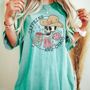 Pu&ograve; includere: Una maglietta blu turchese con una grafica di uno scheletro che indossa un cappello da cowboy e tiene una tazza di caff&egrave; freddo. Il testo "Caffeine and Chaos" &egrave; stampato sotto lo scheletro.