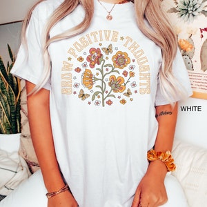 Op de afbeelding: Wit T-shirt met een bloemenontwerp en de tekst "Grow Positive Thoughts" in een cirkel rond de bloemen.