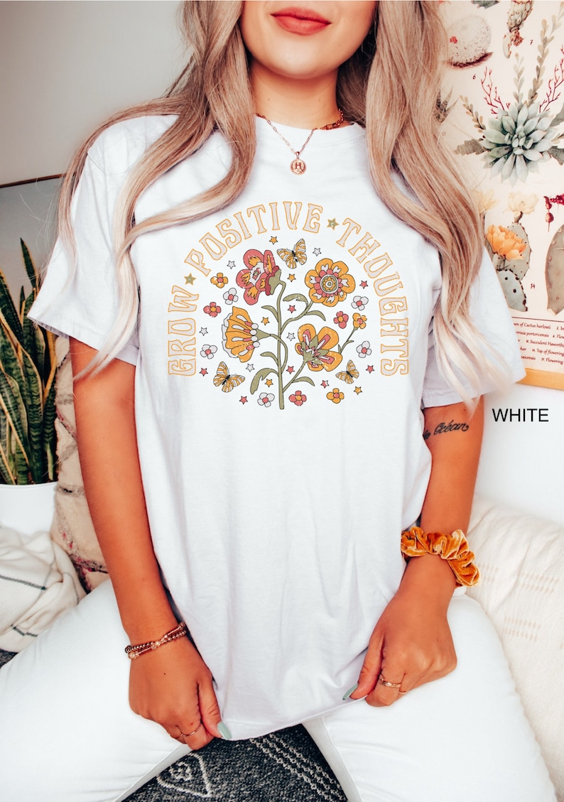 Op de afbeelding: Wit T-shirt met een bloemenontwerp en de tekst "Grow Positive Thoughts" in een cirkel rond de bloemen.