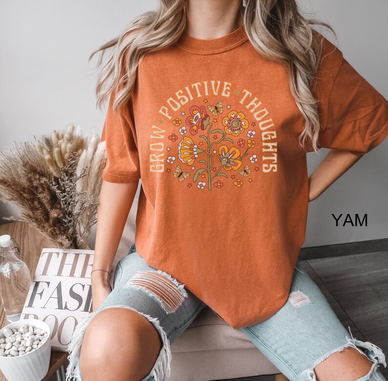Op de afbeelding: Een oranje T-shirt met een bloemenontwerp en de tekst "Grow Positive Thoughts".