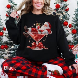 May include: Black sweatshirt featuring a festive Santa Claus illustration relaxing in a cocktail glass filled with red liquid. The design includes a red bow and sparkling accents, perfect for the holiday season.
