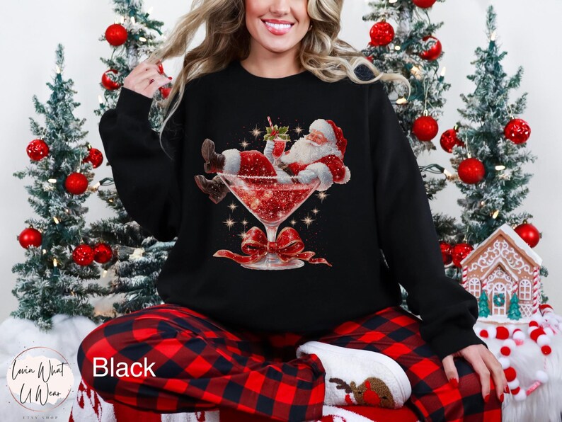 May include: Black sweatshirt featuring a festive Santa Claus illustration relaxing in a cocktail glass filled with red liquid. The design includes a red bow and sparkling accents, perfect for the holiday season.