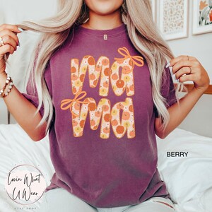 May include: Berry-colored t-shirt with the word "Mama" in large, orange letters filled with a pumpkin pattern. The word "Mama" has orange bow accents. The shirt is a comfortable, casual style.