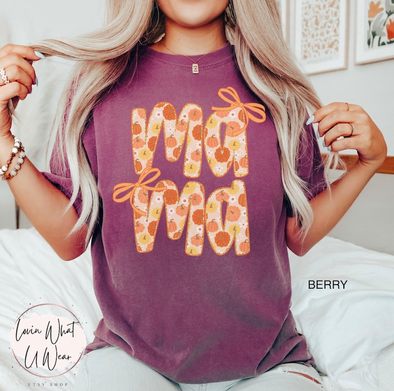 May include: Berry-colored t-shirt with the word "Mama" in large, orange letters filled with a pumpkin pattern. The word "Mama" has orange bow accents. The shirt is a comfortable, casual style.