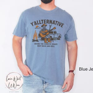 May include: A blue jean-colored t-shirt with a graphic that reads "Y'ALLTERNATIVE" above an illustration of a skeleton playing a guitar next to a campfire and a truck. The text "WHERE THE COUNTRY ROADS MEET ROCK AND ROLL" is below the illustration.