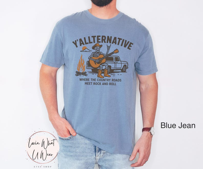 May include: A blue jean-colored t-shirt with a graphic that reads "Y'ALLTERNATIVE" above an illustration of a skeleton playing a guitar next to a campfire and a truck. The text "WHERE THE COUNTRY ROADS MEET ROCK AND ROLL" is below the illustration.