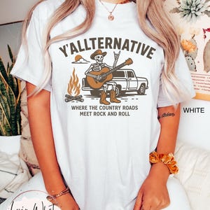 May include: White t-shirt with the text "Y'ALLTERNATIVE" and a graphic of a skeleton playing a guitar in front of a truck and campfire. The phrase "WHERE THE COUNTRY ROADS MEET ROCK AND ROLL" is below the graphic.