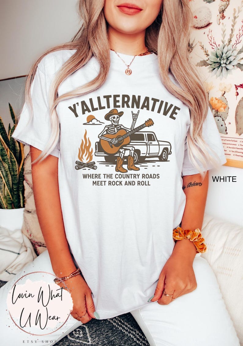 May include: White t-shirt with the text "Y'ALLTERNATIVE" and a graphic of a skeleton playing a guitar in front of a truck and campfire. The phrase "WHERE THE COUNTRY ROADS MEET ROCK AND ROLL" is below the graphic.