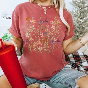 May include: A woman wearing a maroon t-shirt with a floral print. The shirt has a relaxed fit and a crew neckline. The woman is sitting on a white and red plaid blanket.