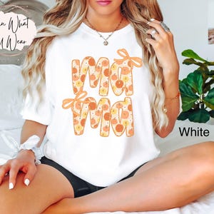 May include: White t-shirt with the word "Mama" in orange letters filled with a pumpkin pattern. The word "Mama" has orange bows on top. The word "White" is printed on the bottom right of the shirt.