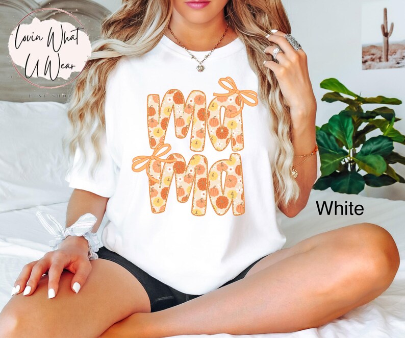 May include: White t-shirt with the word "Mama" in orange letters filled with a pumpkin pattern. The word "Mama" has orange bows on top. The word "White" is printed on the bottom right of the shirt.