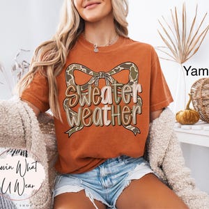 May include: Burnt orange t-shirt with the text "Sweater Weather" in a decorative font, accented by a bow with a camouflage pattern. The model is wearing denim shorts and a cream-colored cardigan.