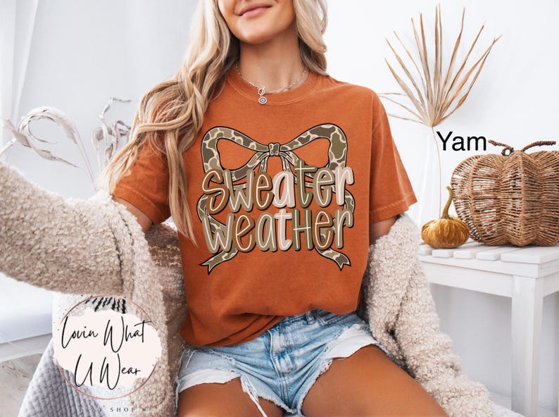 May include: Burnt orange t-shirt with the text "Sweater Weather" in a decorative font, accented by a bow with a camouflage pattern. The model is wearing denim shorts and a cream-colored cardigan.