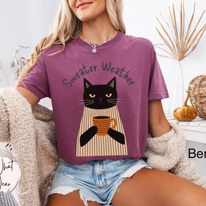 May include: Berry-colored t-shirt with the text "Sweater Weather" above a black cat illustration holding a coffee mug. The cat wears a striped apron. The shirt is paired with denim shorts.