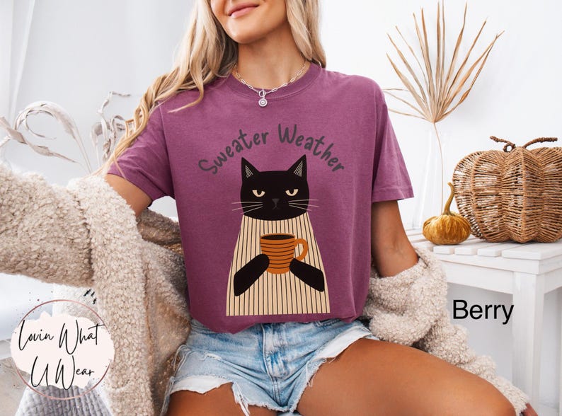 May include: Berry-colored t-shirt with the text "Sweater Weather" above a black cat illustration holding a coffee mug. The cat wears a striped apron. The shirt is paired with denim shorts.