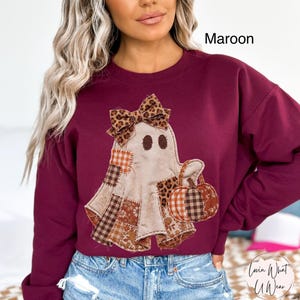 Op de afbeelding: Een kastanjebruin sweatshirt met lange mouwen en een centrale afbeelding van een geest met een strik met luipaardprint. De geest is versierd met diverse patchworkpatronen in bruin-, oranje- en wittinten en houdt een kleine pompoen vast.