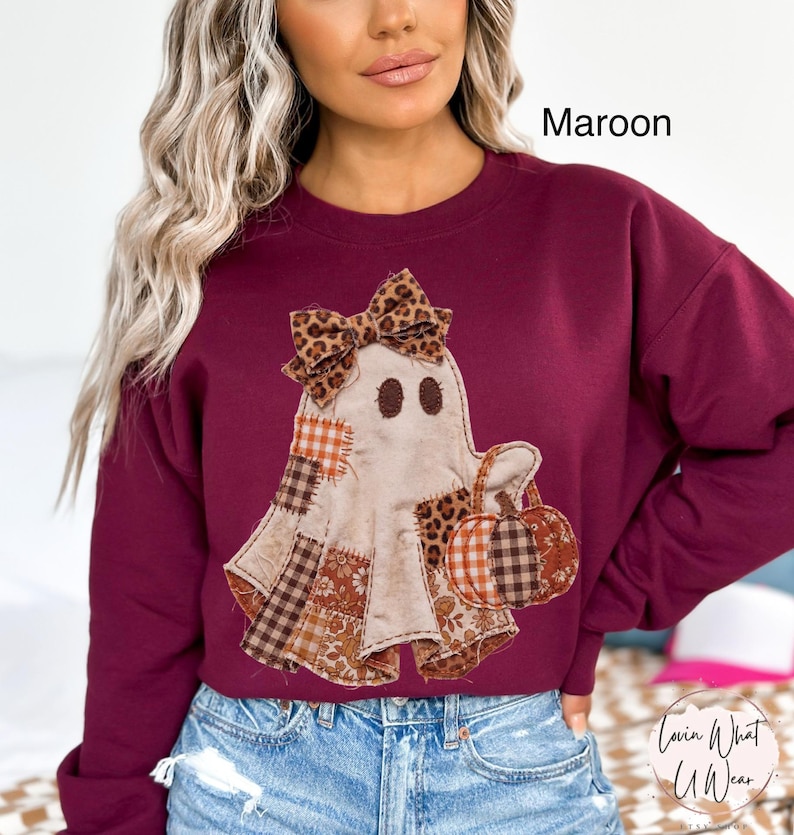 Op de afbeelding: Een kastanjebruin sweatshirt met lange mouwen en een centrale afbeelding van een geest met een strik met luipaardprint. De geest is versierd met diverse patchworkpatronen in bruin-, oranje- en wittinten en houdt een kleine pompoen vast.