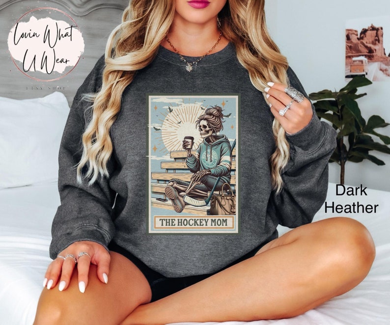 May include: A dark heather grey sweatshirt featuring a graphic print of a skeleton holding a coffee cup and a hockey stick. The graphic also includes the text "The Hockey Mom".