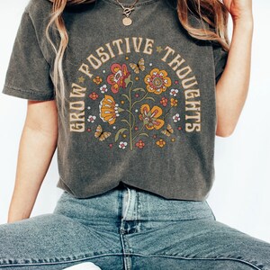 Op de afbeelding: Een donkergrijs T-shirt met een bloemenmotief en de tekst "Grow Positive Thoughts" in een cirkel rond het ontwerp.