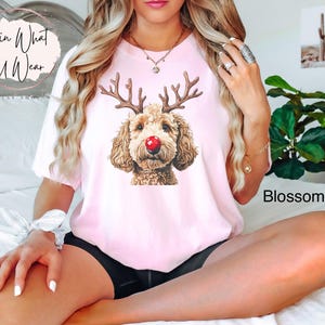 K&ouml;nnte beinhalten: Ein pinkfarbenes T-Shirt mit einem Aufdruck eines braunen und wei&szlig;en Hundes, der eine rote Nase und Rentiergeweih tr&auml;gt. Der Text "Lovin What U Wear" befindet sich in der oberen linken Ecke des Shirts.