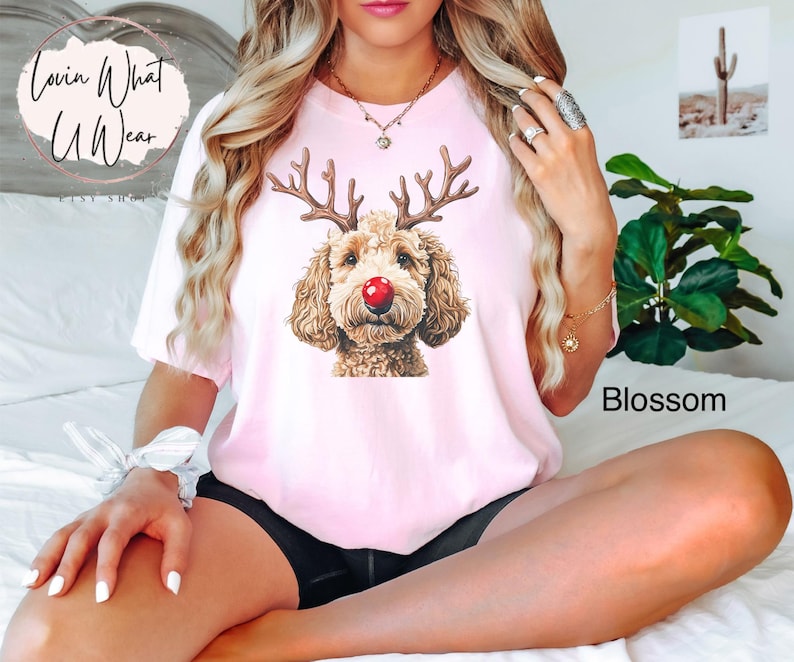 K&ouml;nnte beinhalten: Ein pinkfarbenes T-Shirt mit einem Aufdruck eines braunen und wei&szlig;en Hundes, der eine rote Nase und Rentiergeweih tr&auml;gt. Der Text "Lovin What U Wear" befindet sich in der oberen linken Ecke des Shirts.
