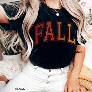 May include: A black t-shirt with a vintage-style "FALL" graphic in shades of orange, red, and yellow. The shirt is worn by a person with long blonde hair and white jeans.