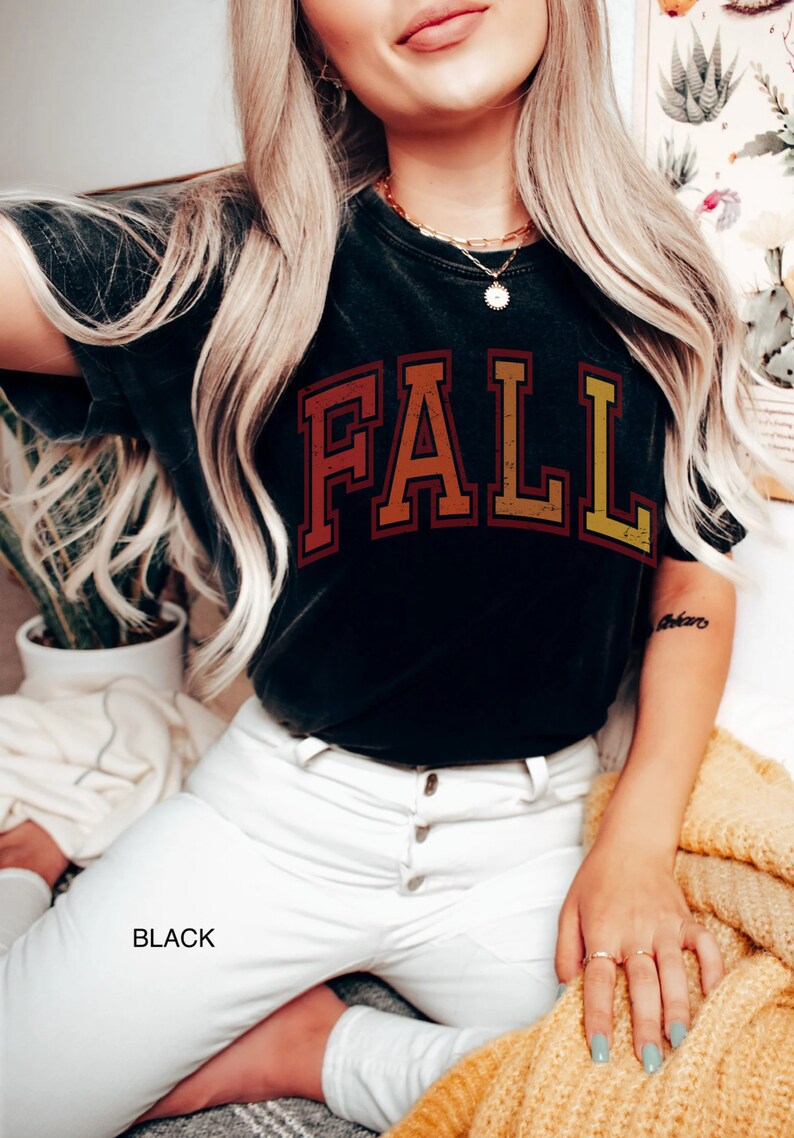 May include: A black t-shirt with a vintage-style "FALL" graphic in shades of orange, red, and yellow. The shirt is worn by a person with long blonde hair and white jeans.