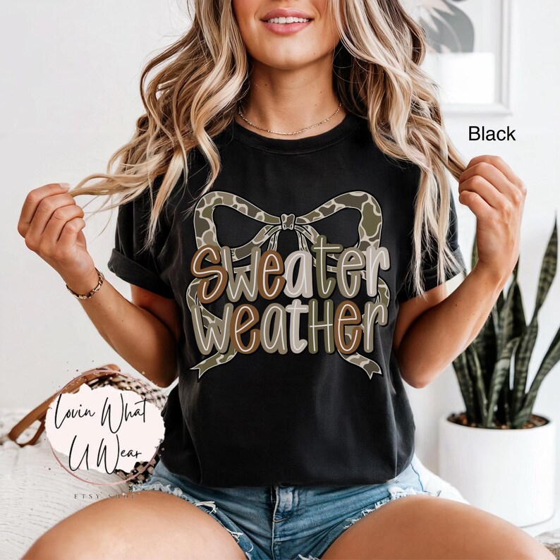 May include: Black t-shirt with the text "Sweater Weather" in a decorative font, accented by a camouflage bow. The shirt is a casual style, suitable for everyday wear.