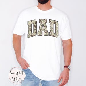 May include: White t-shirt with the word "DAD" in a camouflage pattern across the chest. The letters are outlined in a darker color, creating a bold contrast against the white fabric. The shirt is a casual style.