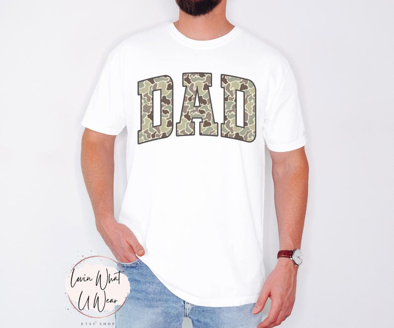 May include: White t-shirt with the word "DAD" in a camouflage pattern across the chest. The letters are outlined in a darker color, creating a bold contrast against the white fabric. The shirt is a casual style.