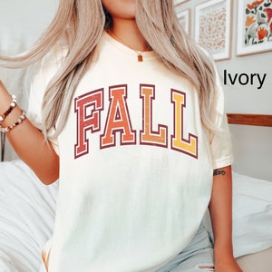 May include: A white t-shirt with a vintage-style "FALL" graphic in red, orange, and yellow. The shirt is shown on a model wearing blue jeans.