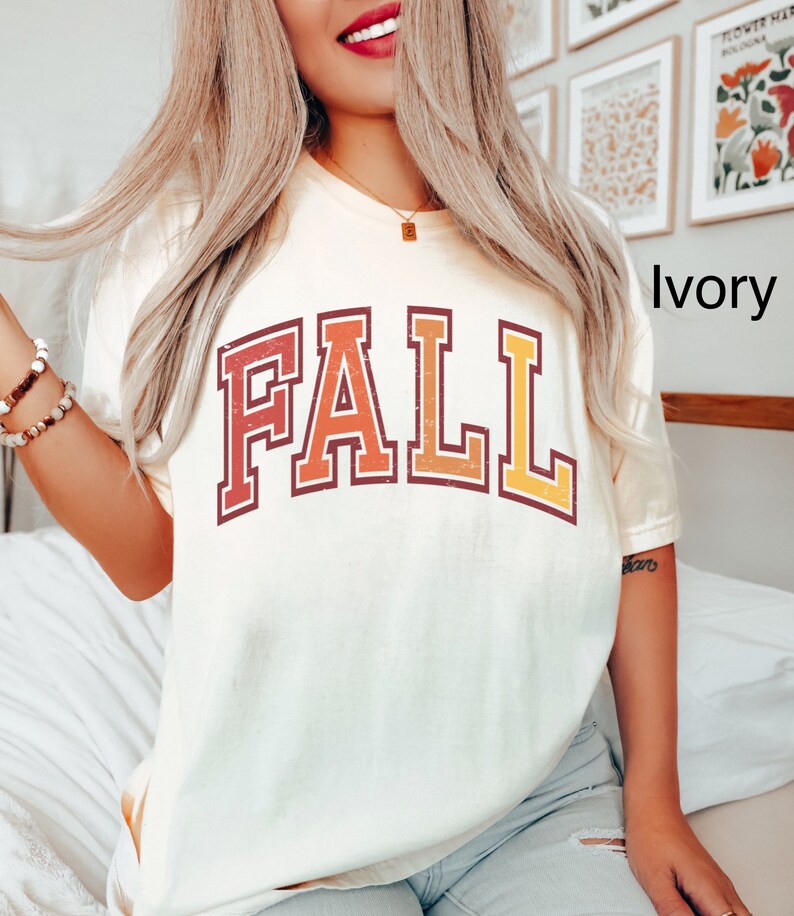May include: A white t-shirt with a vintage-style "FALL" graphic in red, orange, and yellow. The shirt is shown on a model wearing blue jeans.