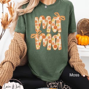 May include: A moss green t-shirt with the word "Mama" in orange letters filled with pumpkin patterns. The shirt has orange bows above the letters. The text "Moss" is visible on the lower right. The brand "Lovin What U Wear" is visible on the lower left.