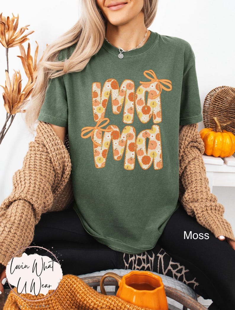 May include: A moss green t-shirt with the word "Mama" in orange letters filled with pumpkin patterns. The shirt has orange bows above the letters. The text "Moss" is visible on the lower right. The brand "Lovin What U Wear" is visible on the lower left.