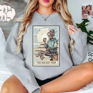 May include: A grey sweatshirt featuring a graphic of a skeleton woman sitting on a bench holding a coffee cup. The graphic is in a vintage style with a light blue background and the text "The Hockey Mom".
