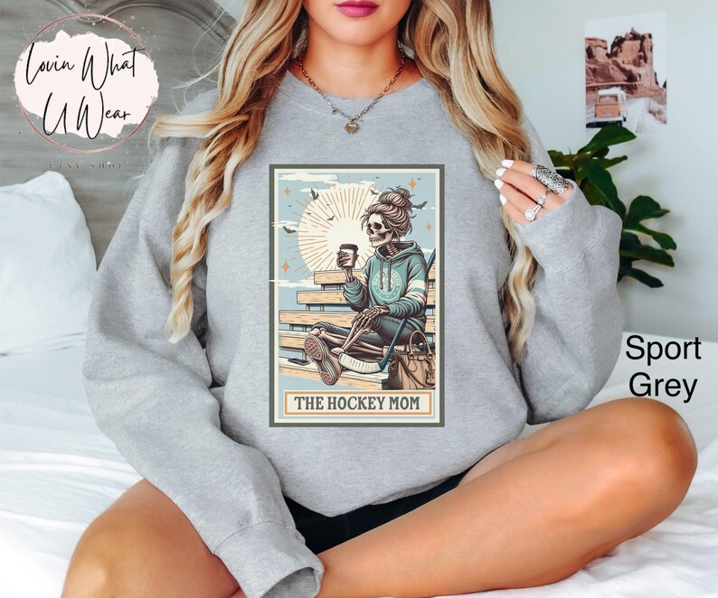 May include: A grey sweatshirt featuring a graphic of a skeleton woman sitting on a bench holding a coffee cup. The graphic is in a vintage style with a light blue background and the text "The Hockey Mom".