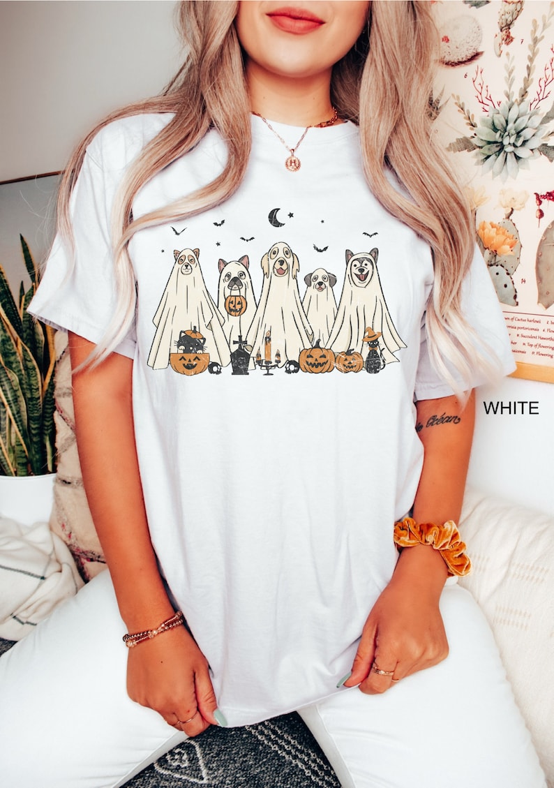 Peut inclure: T-shirt blanc avec un motif de cinq chiens habill&eacute;s en fant&ocirc;mes tenant des citrouilles et des bonbons. Les chiens portent des costumes de fant&ocirc;mes avec une lune croissante et des chauves-souris en arri&egrave;re-plan.
