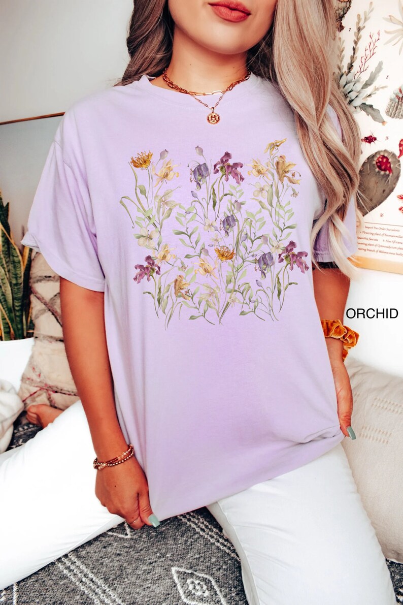 May include: A light purple t-shirt with a floral print featuring yellow, purple, and green flowers. The shirt has a relaxed fit and short sleeves.