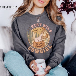May include: Dark heather grey sweatshirt with a graphic print of a woman in cowboy boots sitting in a jack-o-lantern. The graphic also includes the text "Stay Wild Cowgirl".