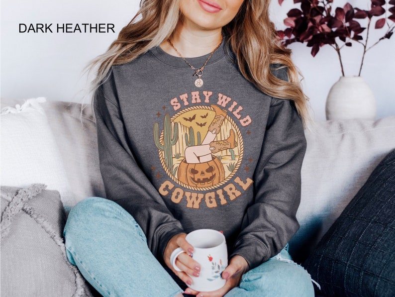 May include: Dark heather grey sweatshirt with a graphic print of a woman in cowboy boots sitting in a jack-o-lantern. The graphic also includes the text "Stay Wild Cowgirl".
