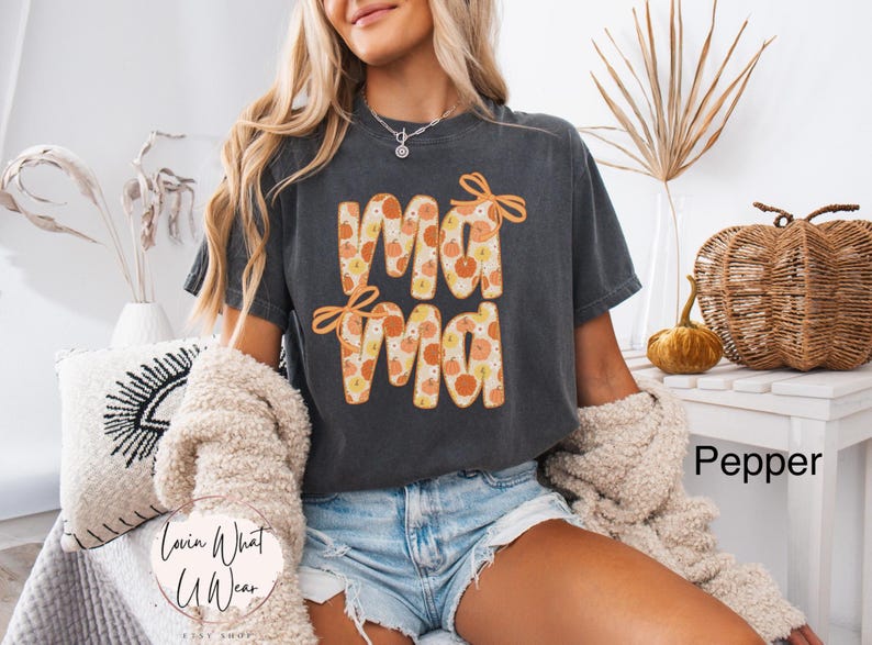 May include: A dark gray t-shirt with the word "Mama" in orange letters filled with a pumpkin pattern. The shirt has orange bows above the first "Ma" and below the second "Ma". The shirt is part of the Lovin What U Wear Etsy shop.